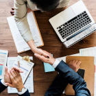 Overhead view of a handshake across a desk with documents and a laptop